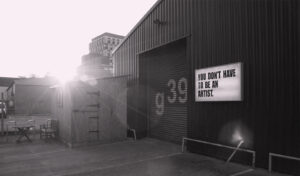 A picture of the front of a metal warehouse with a roller shutter displaying g39. A lightbox to the right of the shutter says 'You Don't Have To Be An Artist'